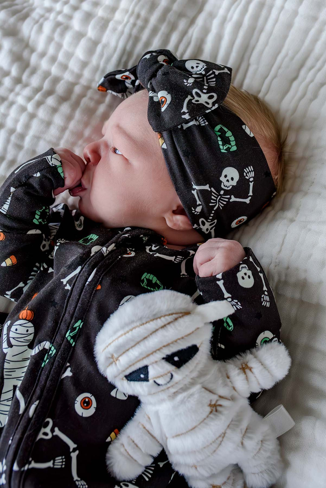 Newborn wearing a Caden Lane black skeleton and mummy print footie with matching bow headband, lying on the coordinating blanket and holding a plush mummy toy.