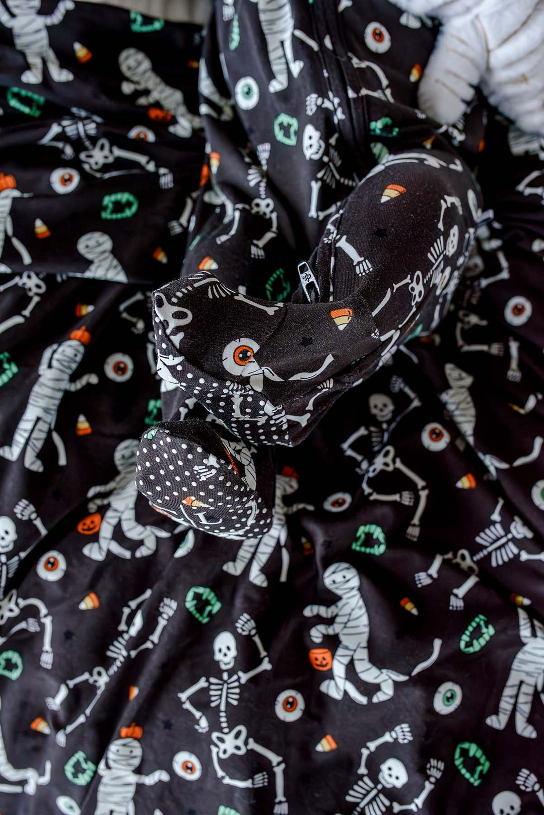 Baby wearing a Caden Lane black skeleton and mummy print footie with matching bow headband, lying on the coordinating blanket and holding a plush mummy toy.