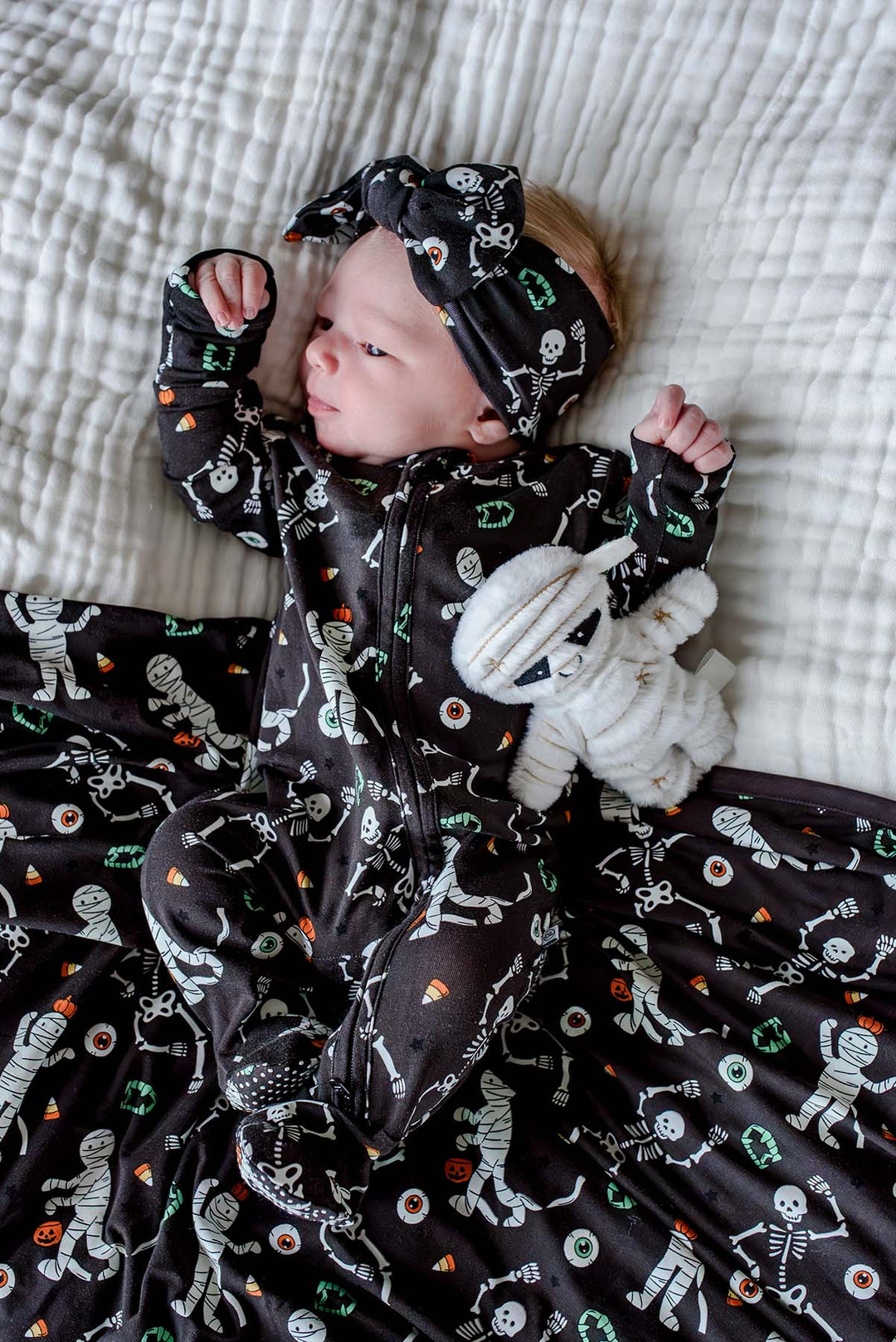 Newborn wearing a Caden Lane black skeleton and mummy print footie with matching bow headband, lying on the coordinating blanket and holding a plush mummy toy.