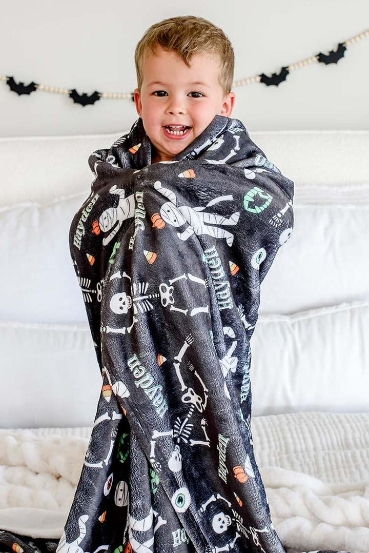 A young boy smiling while wrapped in a dark gray Halloween-themed blanket with white and orange graphics, sitting on a white bed with pillows and a bat banner in the background.