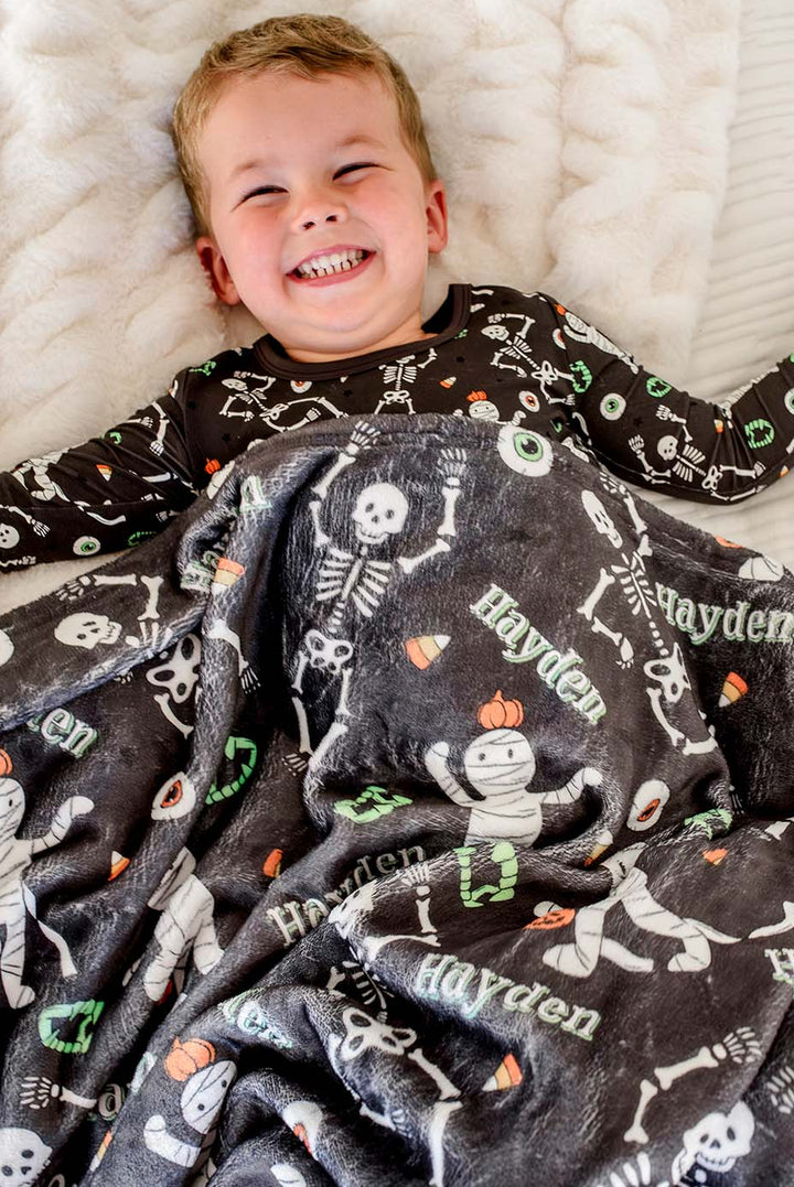 A smiling young boy lies on a white textured blanket, wearing a black long-sleeve pajama top with a white skeleton print and a dark gray personalized fleece blanket printed with white skulls, orange pumpkins, and the name 'Hayden'.