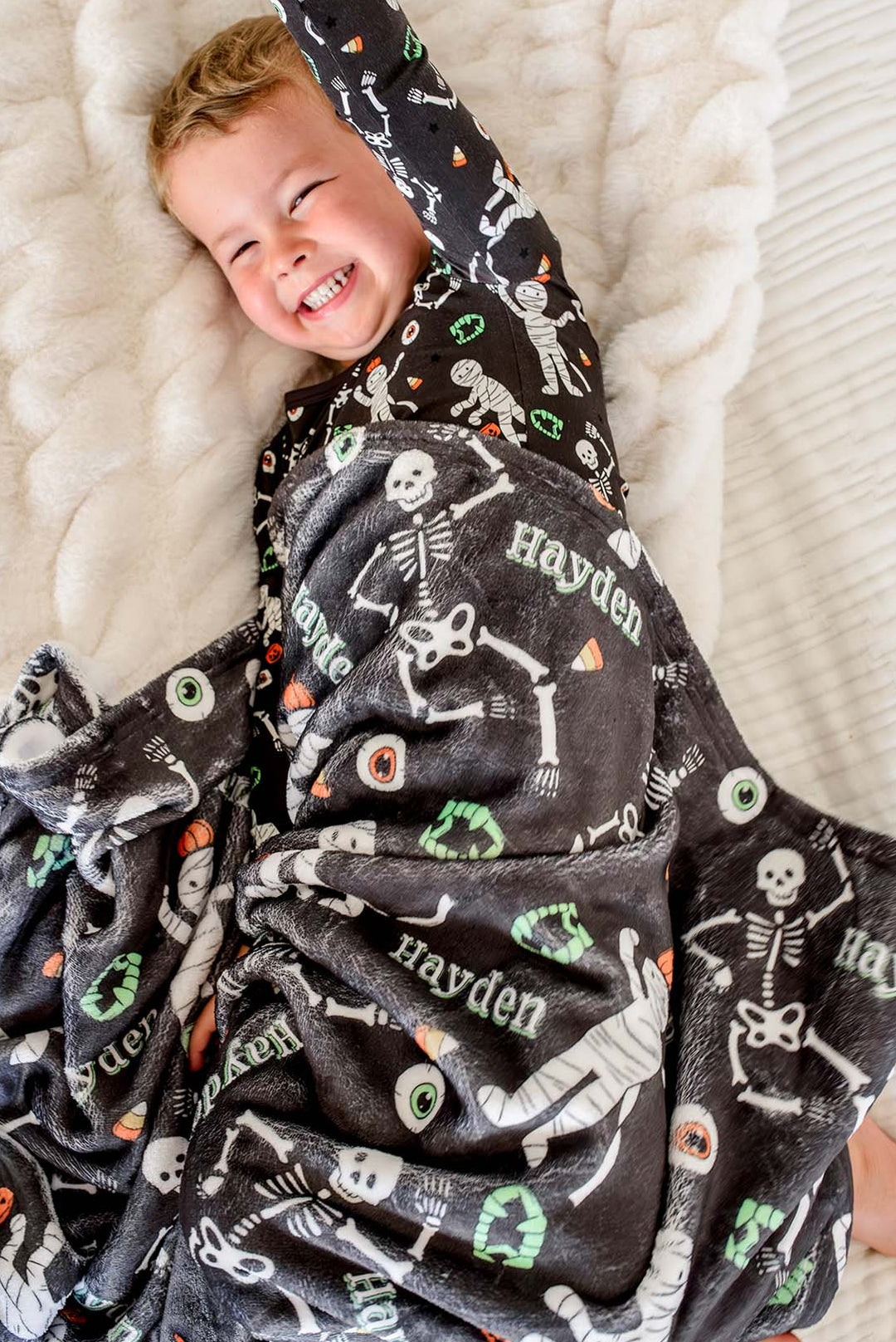 A smiling toddler lying on a white textured blanket, wrapped in a dark gray plush Halloween-themed blanket printed with white skeletons, candy corn, eyeballs, and the name 'Hayden'.