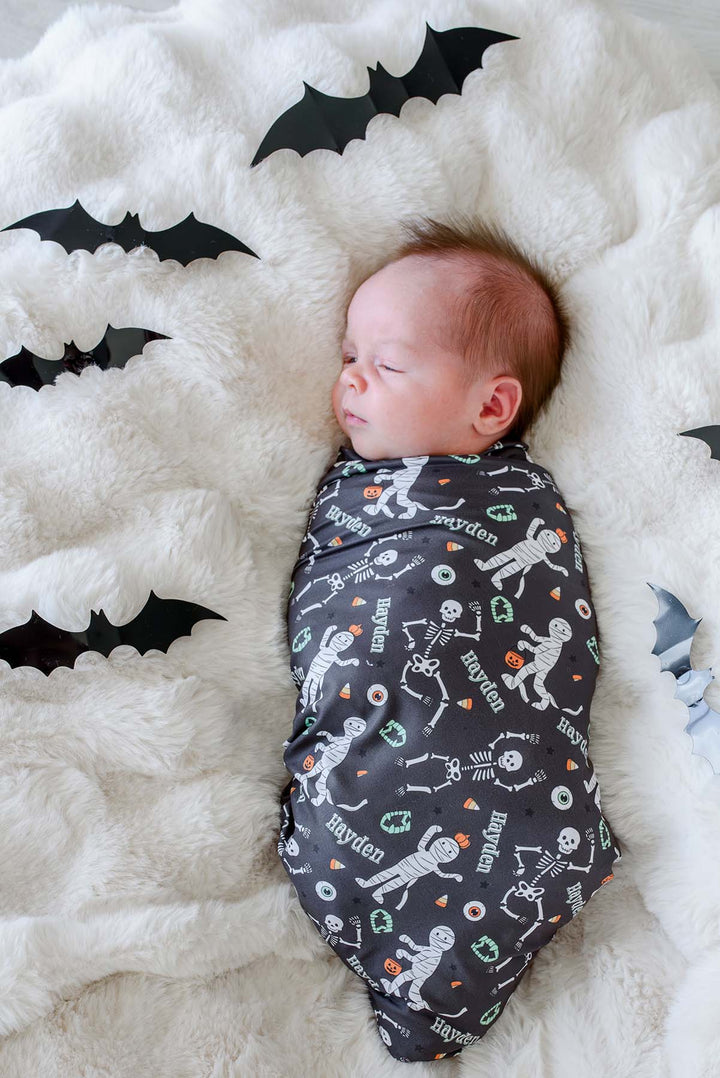 A sleeping newborn baby wrapped in a dark gray Halloween-themed swaddle blanket with skeleton and pumpkin prints, lying on a white fluffy blanket decorated with black bat cutouts.