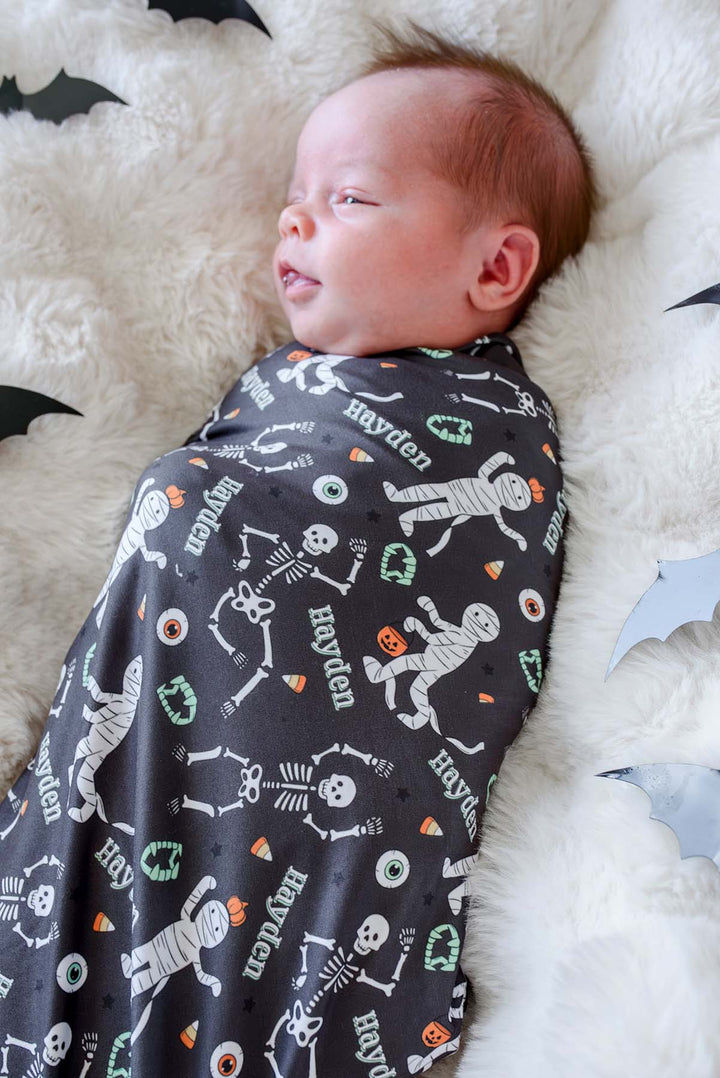 A newborn baby wrapped in a dark gray Halloween-themed swaddle blanket with white skeleton prints, orange pumpkins, and the name 'Hayden' on a fluffy white blanket.