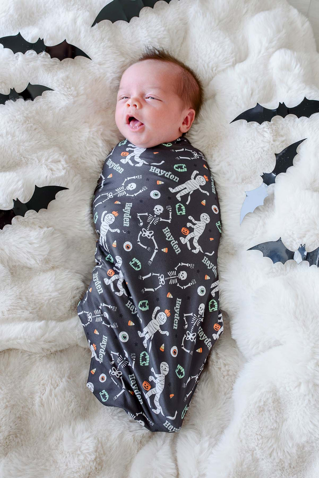 A baby swaddled in a dark gray Halloween-themed fabric blanket personalized with the name 'Hayden' and printed with skeleton and mummy illustrations, lying on a fluffy white blanket surrounded by black bat cutouts.