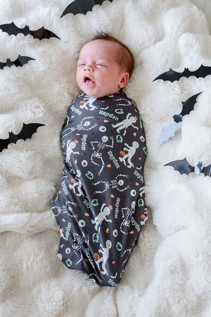 A baby swaddled in a dark gray Halloween-themed fabric blanket personalized with the name 'Hayden' and printed with skeleton and mummy illustrations, lying on a fluffy white blanket surrounded by black bat cutouts.