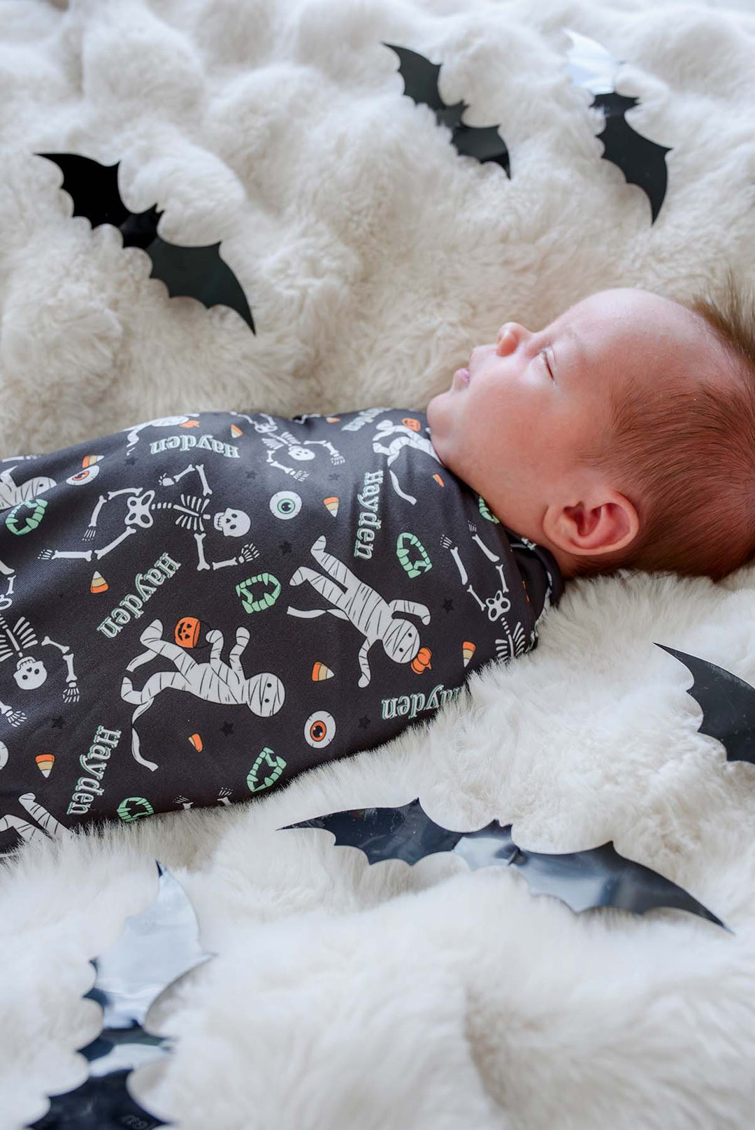 A baby sleeping on a fluffy white blanket, wearing a dark gray Halloween-themed swaddle with skeleton, mummy, and pumpkin prints. Black bat cutouts are scattered around the blanket.