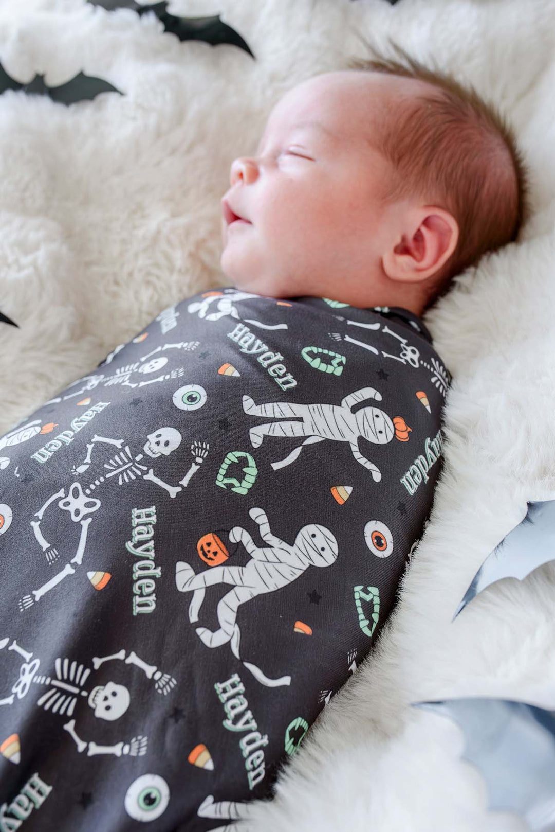 A sleeping newborn baby wrapped in a dark gray Halloween-themed swaddle blanket with white mummies, skeletons, and candy corn, resting on a white faux fur blanket.