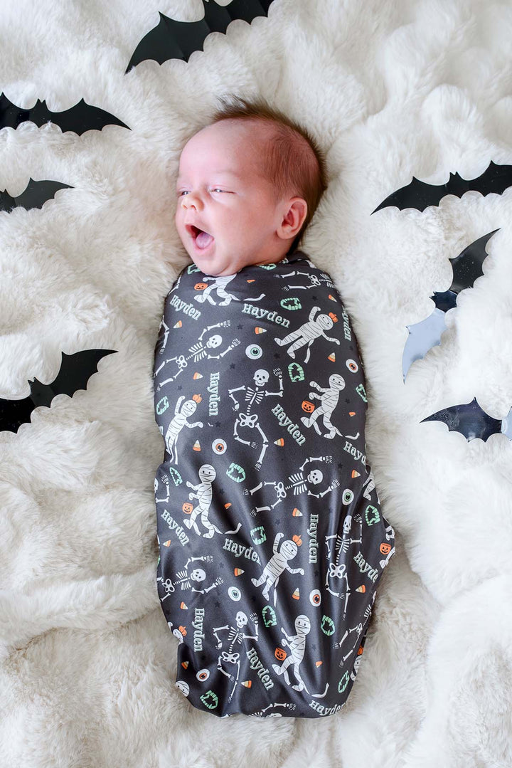 A newborn baby is swaddled in a dark gray Halloween-themed blanket personalized with the name 'Hayden' and printed with white skeleton and mummy graphics, lying on a white fluffy blanket decorated with black bat cutouts.
