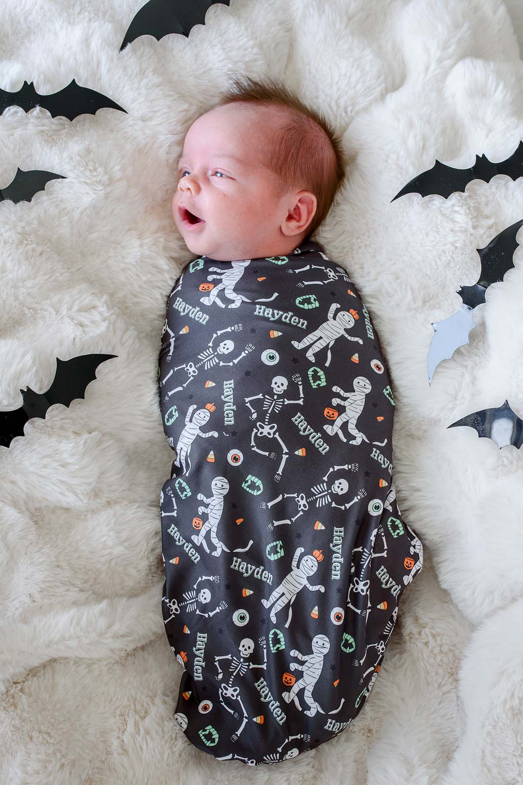 A baby swaddled in a dark gray Halloween-themed blanket with skeleton and pumpkin prints, lying on a white fluffy rug with black bat cutouts.