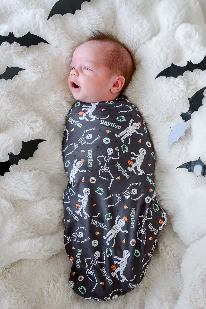 A baby swaddled in a dark gray Halloween-themed blanket with skeleton and pumpkin prints, lying on a white fluffy rug with black bat cutouts.