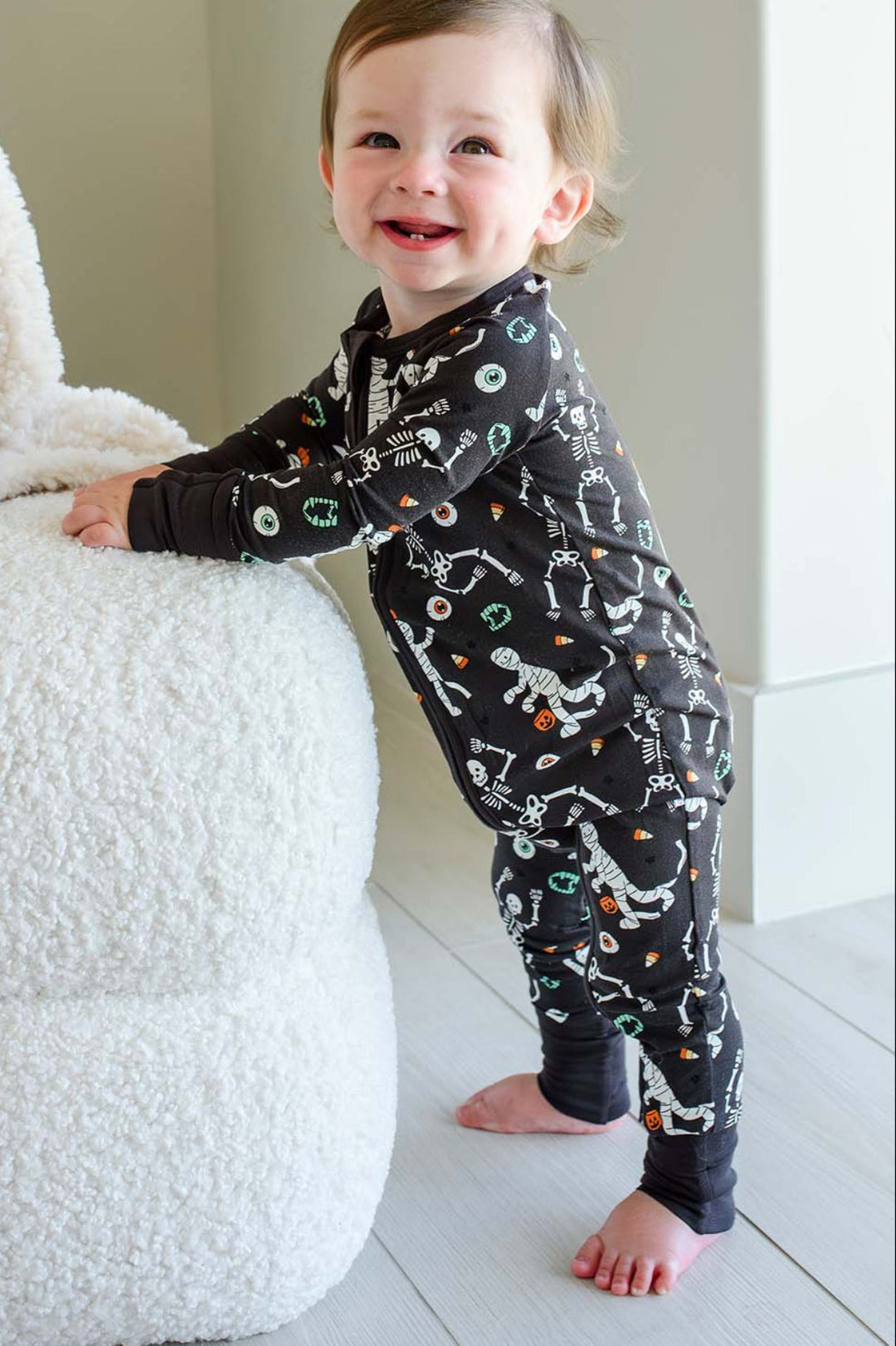 Smiling toddler standing beside a plush white ghost and holding a mummy toy, wearing a Caden Lane black zip-up skeleton and mummy print romper.
