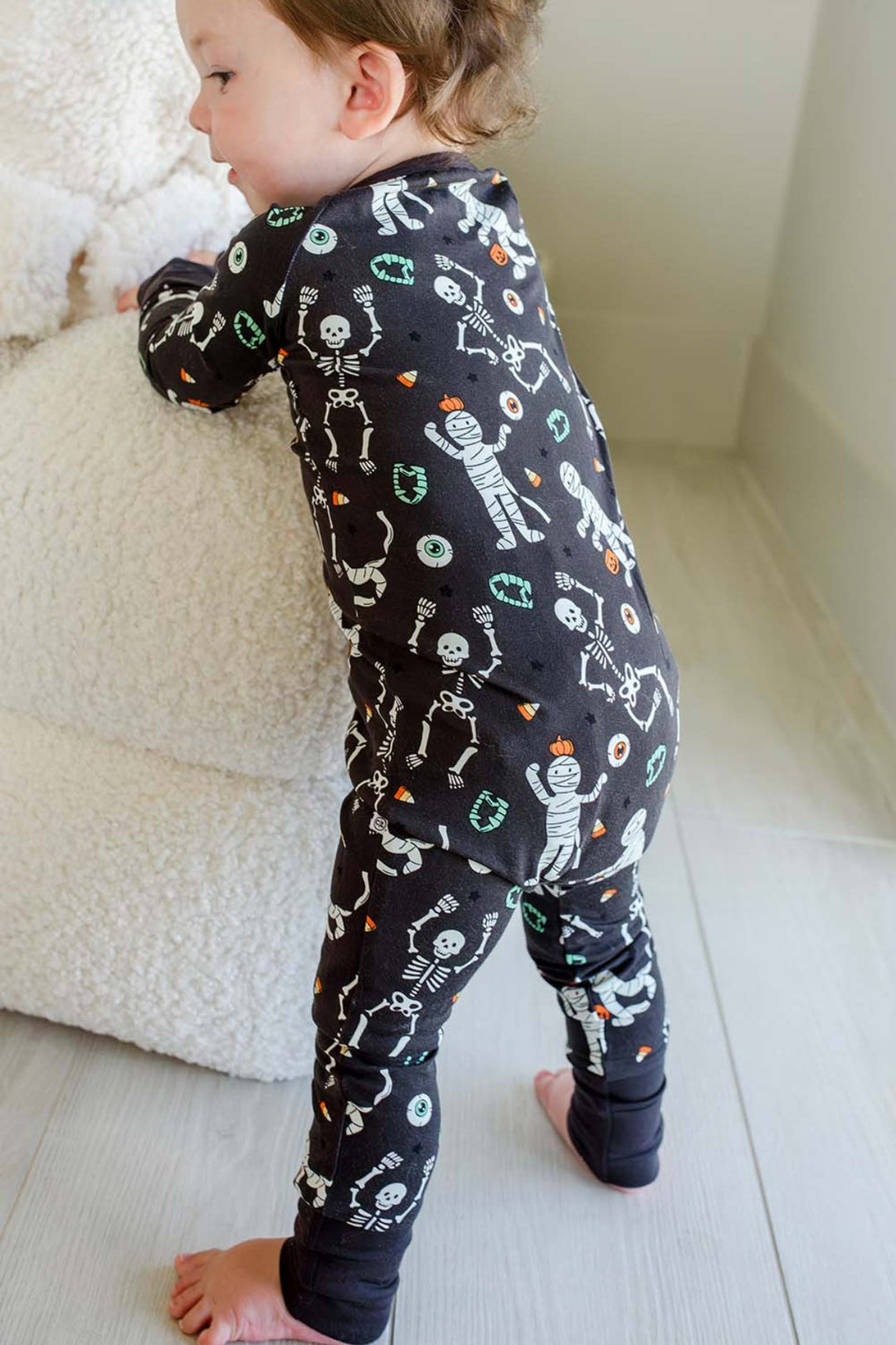 Toddler standing beside a plush white ghost and holding a mummy toy, wearing a Caden Lane black zip-up skeleton and mummy print romper.