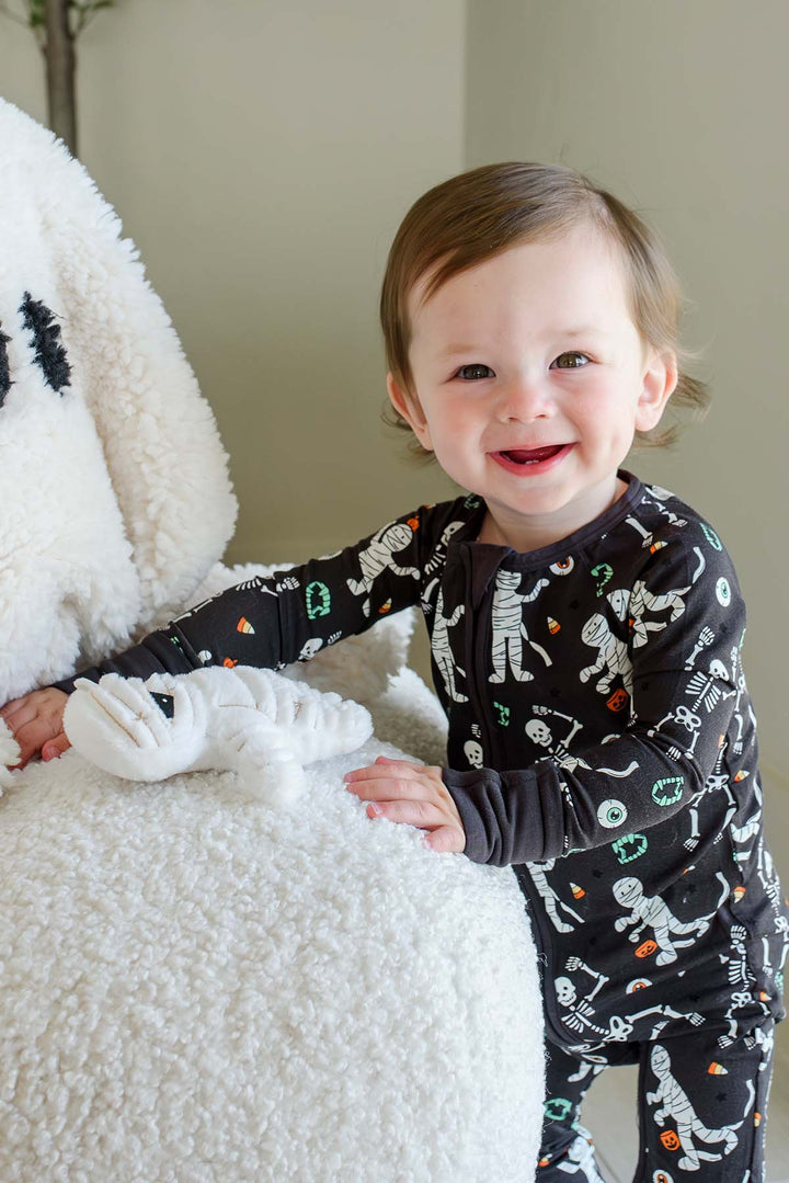 Smiling toddler standing beside a plush white ghost and holding a mummy toy, wearing a Caden Lane black zip-up skeleton and mummy print romper.