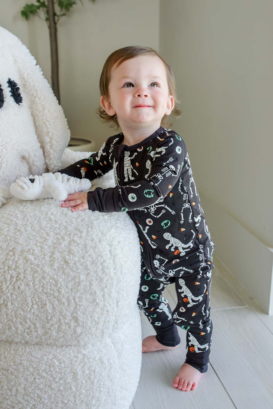 Smiling toddler standing beside a plush white ghost and holding a mummy toy, wearing a Caden Lane black zip-up skeleton and mummy print romper.