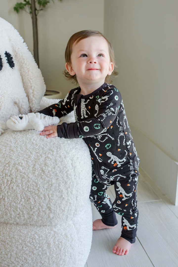 Smiling toddler standing beside a plush white ghost and holding a mummy toy, wearing a Caden Lane black zip-up skeleton and mummy print romper.