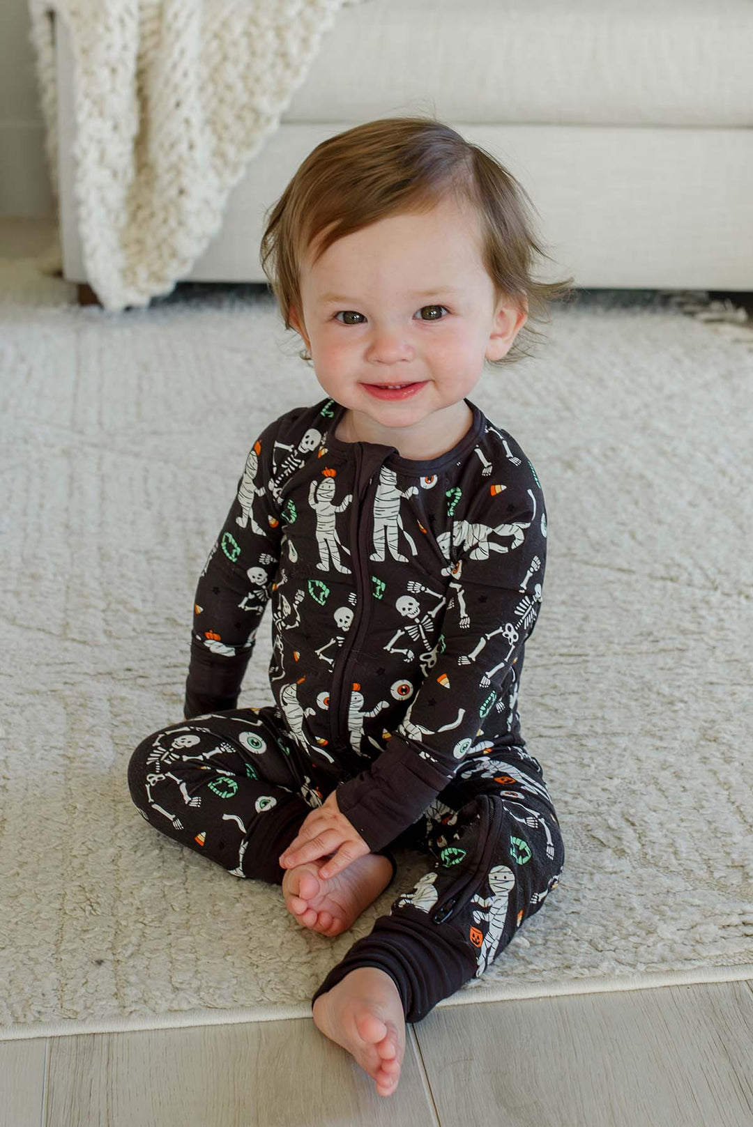 Smiling toddler wearing a Caden Lane black zip-up skeleton and mummy print romper.