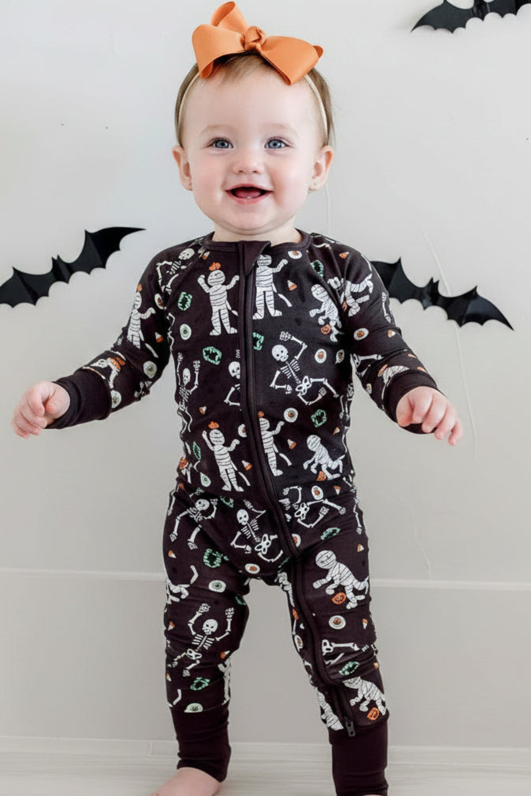 Smiling baby standing and wearing a Caden Lane black zip-up skeleton and mummy print romper with an orange bow, against a bat-decorated Halloween backdrop.