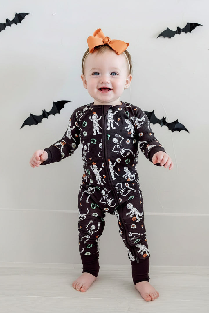 Smiling baby standing and wearing a Caden Lane black zip-up skeleton and mummy print romper with an orange bow, against a bat-decorated Halloween backdrop.