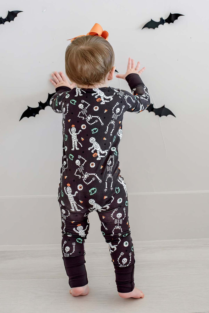 Smiling baby standing and wearing a Caden Lane black zip-up skeleton and mummy print romper with an orange bow, against a bat-decorated Halloween backdrop.