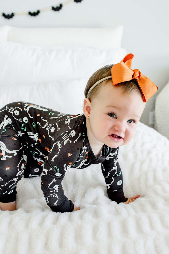 Baby wearing a Caden Lane black zip-up skeleton and mummy print romper with an orange bow, against bed with white bedding
