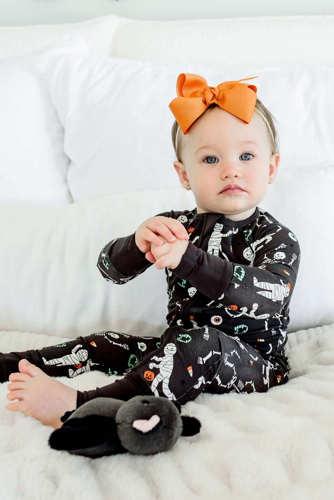 Sitting baby wearing a Caden Lane black zip-up skeleton and mummy print romper with an orange bow, against a bed with white bedding