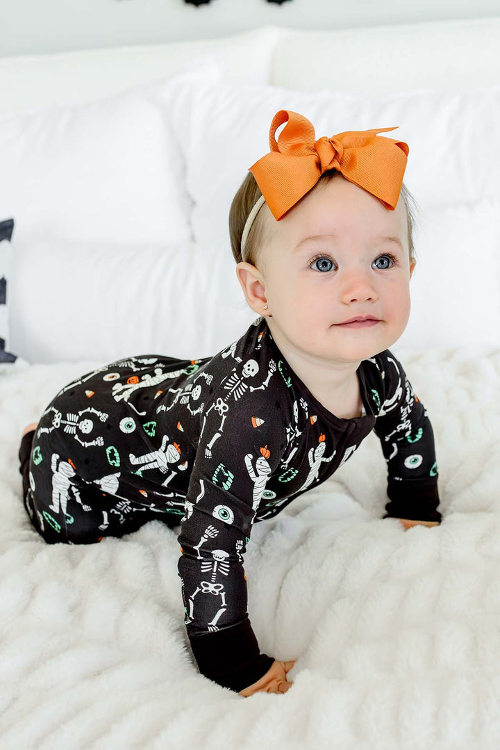 Smiling baby wearing a Caden Lane black zip-up skeleton and mummy print romper with an orange bow, against a bed with white bedding