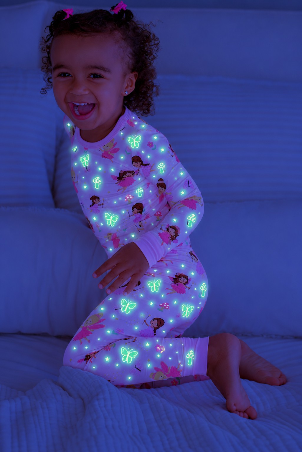 Laughing girl kneeling on a bed, wearing a glowing Caden Lane pink bamboo two-piece pajama set in a Fairy Wishes print featuring fairies, butterflies, and stars.