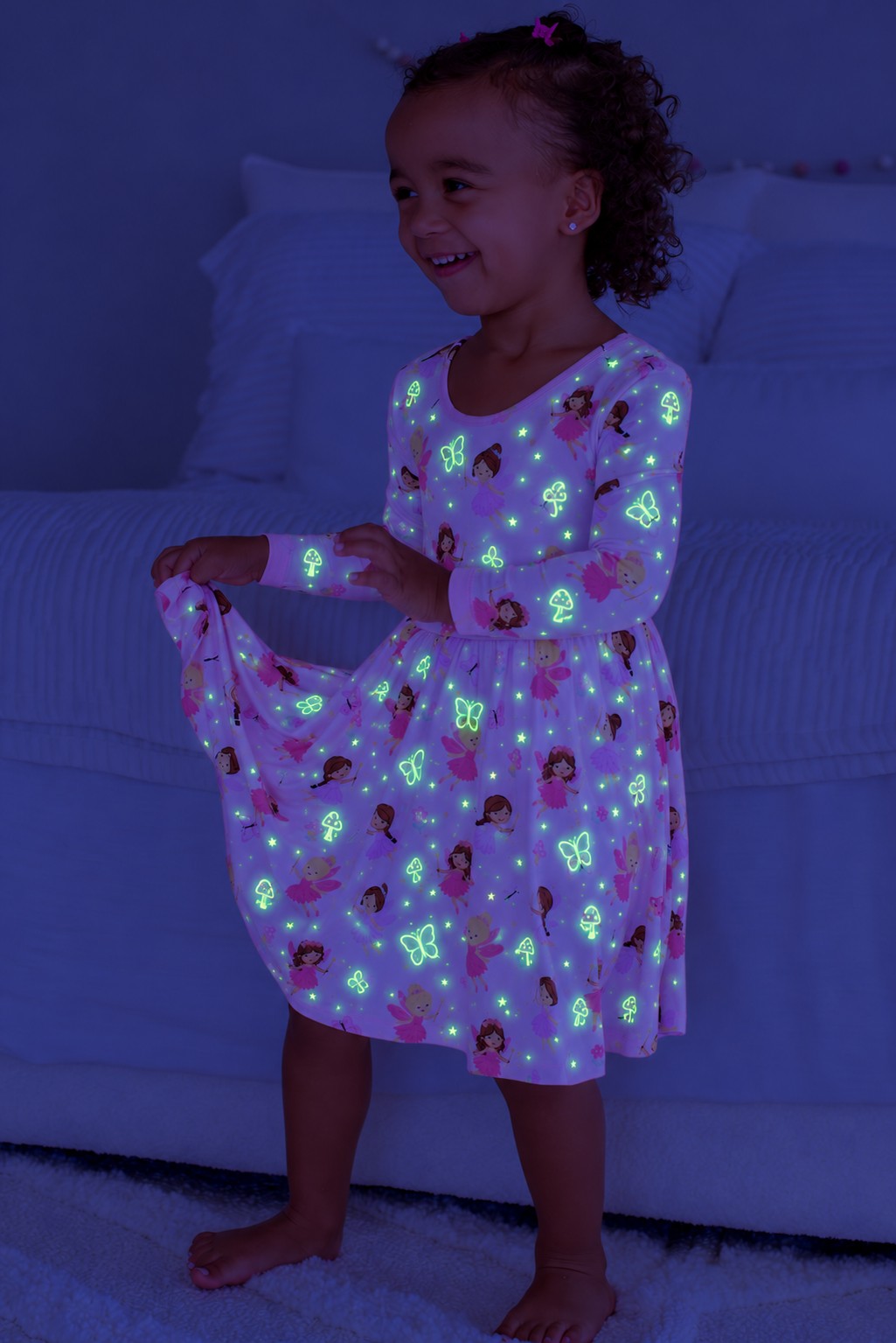 Smiling girl holding out her skirt, wearing a glowing Caden Lane pink bamboo long sleeve twirl dress in a Fairy Wishes print featuring fairies and butterflies.