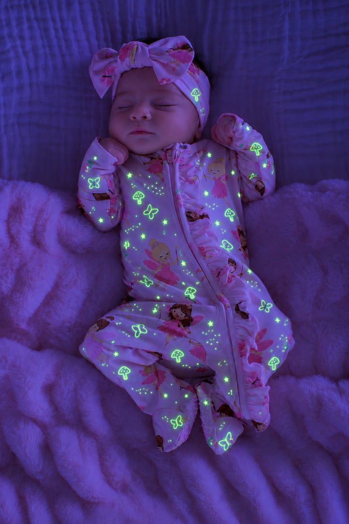 Sleeping newborn on a fluffy blanket, wearing a glowing Caden Lane pink bamboo zipper footie in a Fairy Wishes print featuring fairies and butterflies, paired with a matching bow headwrap.