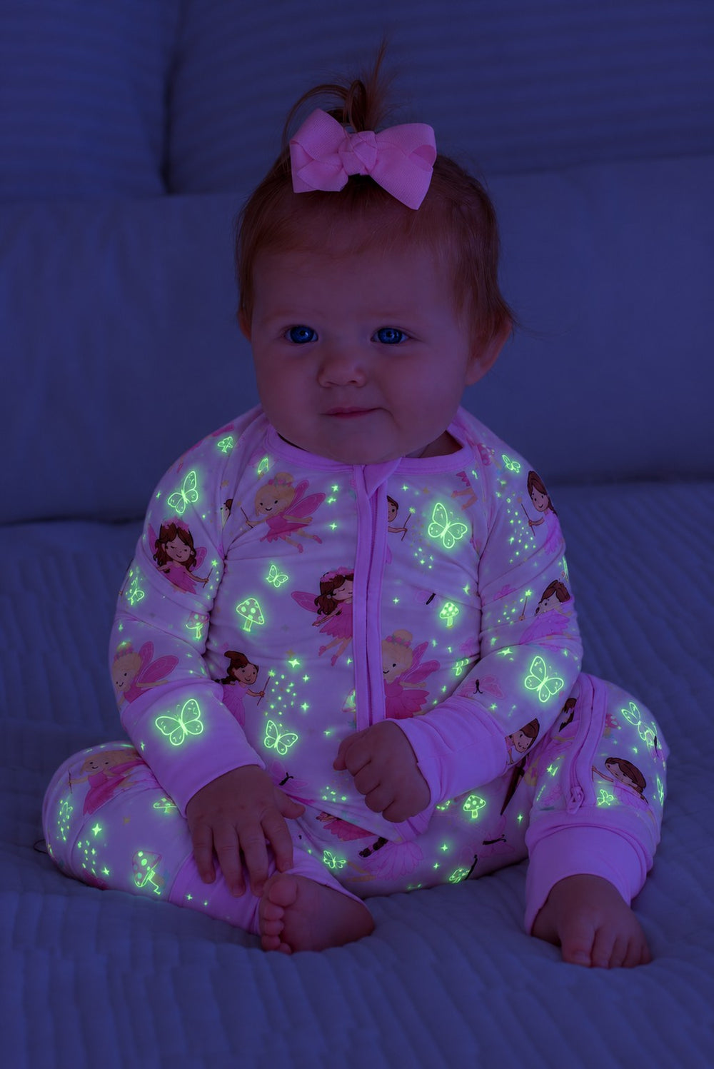 Baby with a pink hair bow sitting on a bed, wearing a glowing Caden Lane pink bamboo convertible zip romper in a Fairy Wishes print featuring fairies and butterflies.