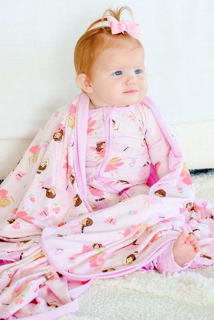 Baby girl sitting wrapped in the Fairy Wishes glow-in-the-dark double-sided bamboo blanket, showing the fairy print and pink trim