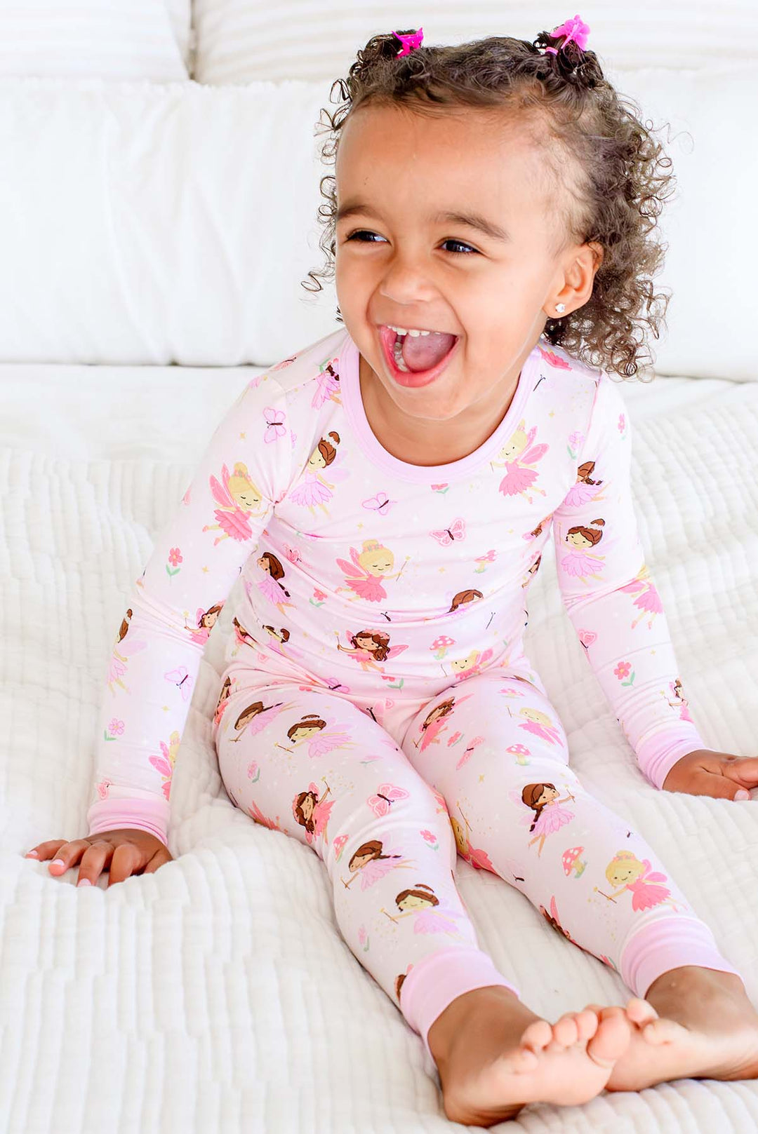 Laughing toddler girl sitting on a bed in the Fairy Wishes glow-in-the-dark two-piece bamboo pajamas with fairy print