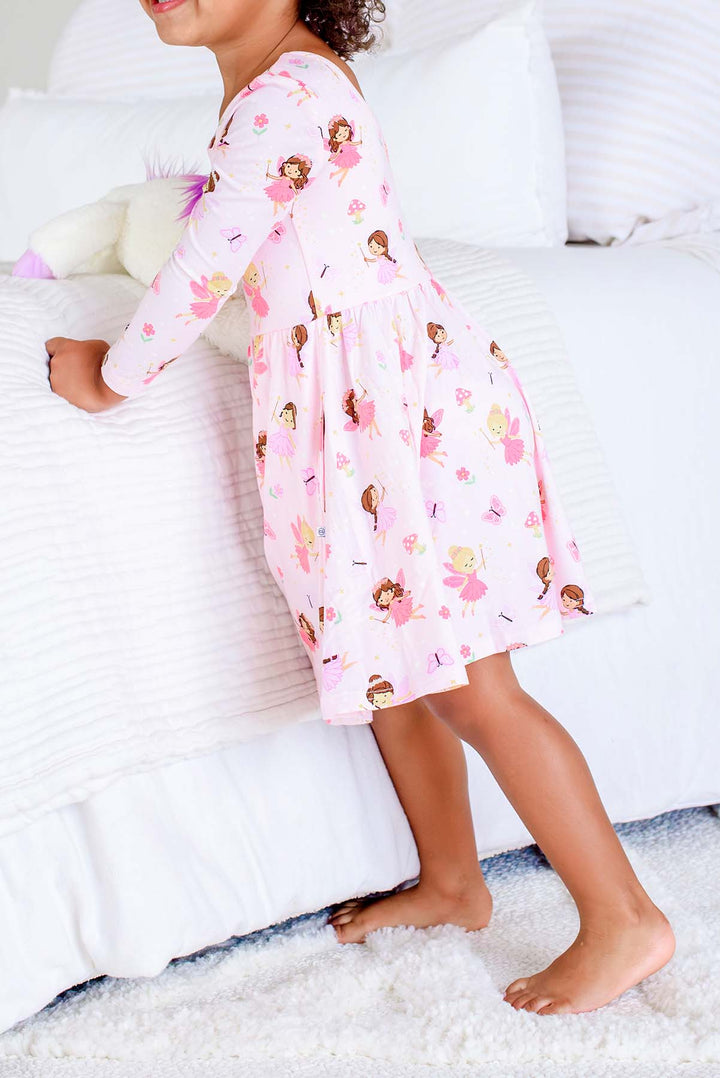 Toddler in the Fairy Wishes long sleeve twirl dress leaning against a white bed, side view of the fairy print dress