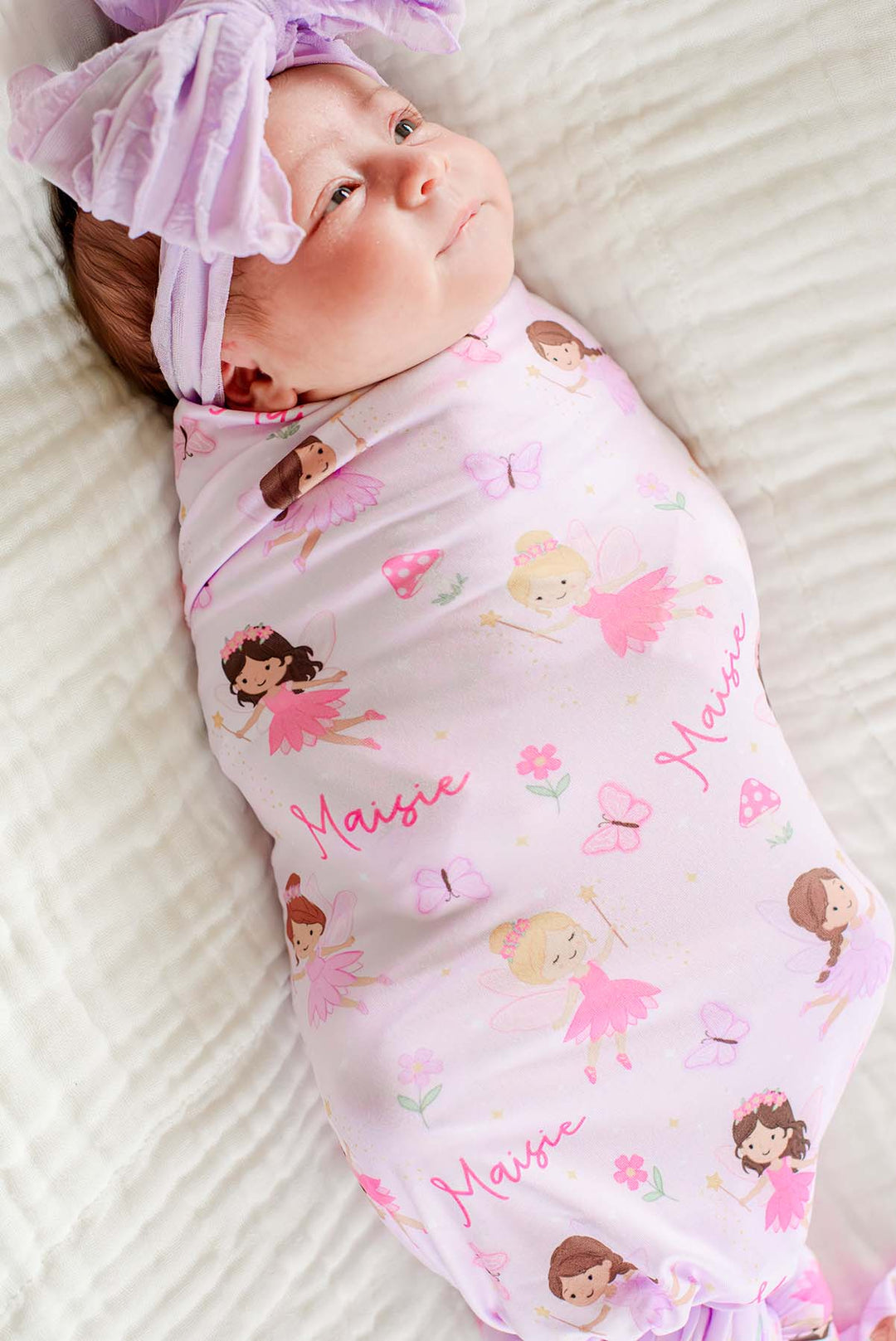 Close-up of a swaddled baby in the Fairy Wishes personalized swaddle with fairies and custom name print, wearing a lavender bow