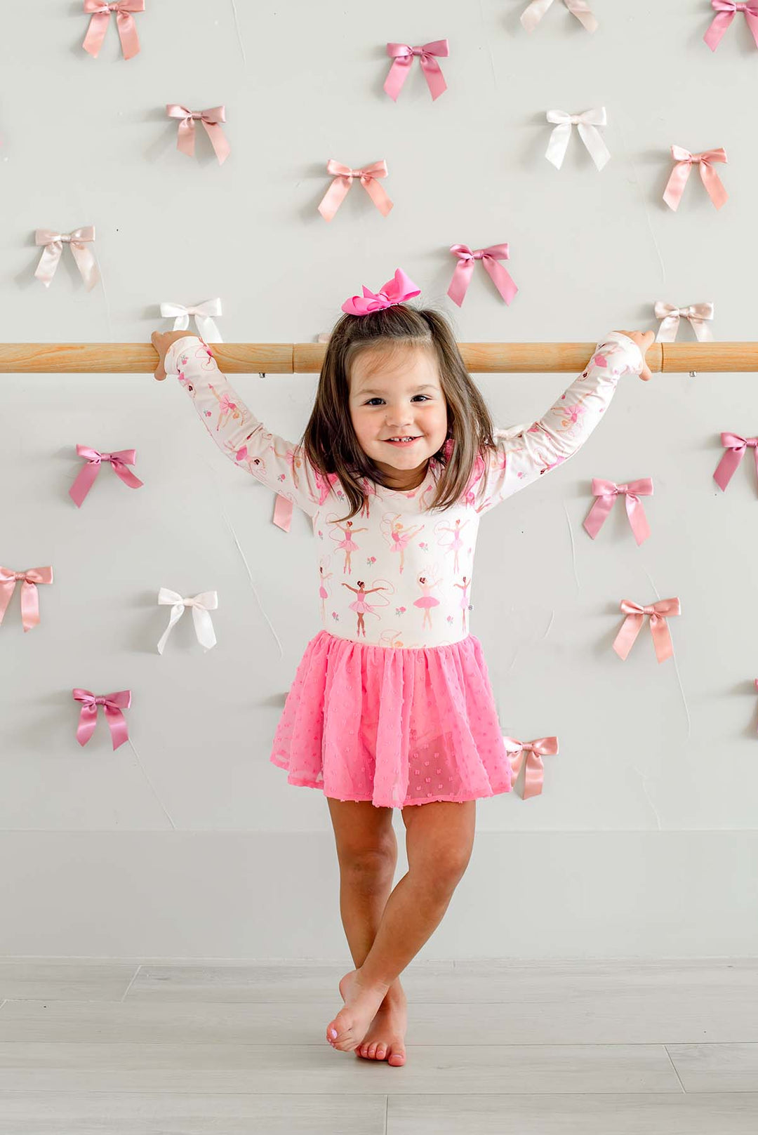 Smiling girl holding a ballet barre with crossed ankles, wearing a Caden Lane long-sleeve leotard in a ballerina print with a pink swiss dot mesh skirt.