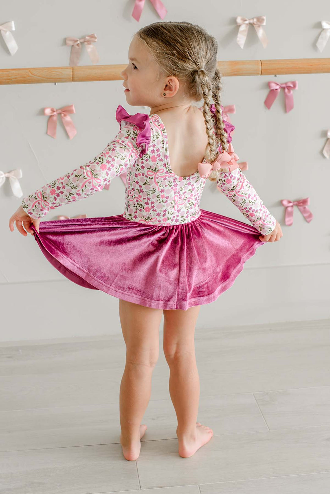 Back view of a girl holding out the skirt of a Caden Lane long-sleeve leotard in a pink floral and bow print with a berry velvet skirt, standing in front of a ballet barre.