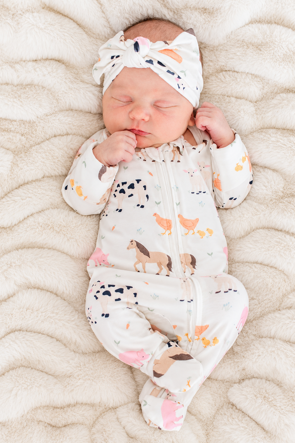 Sleeping newborn in Caden Lane farm animal print zip-up footie pajamas with matching bow headband