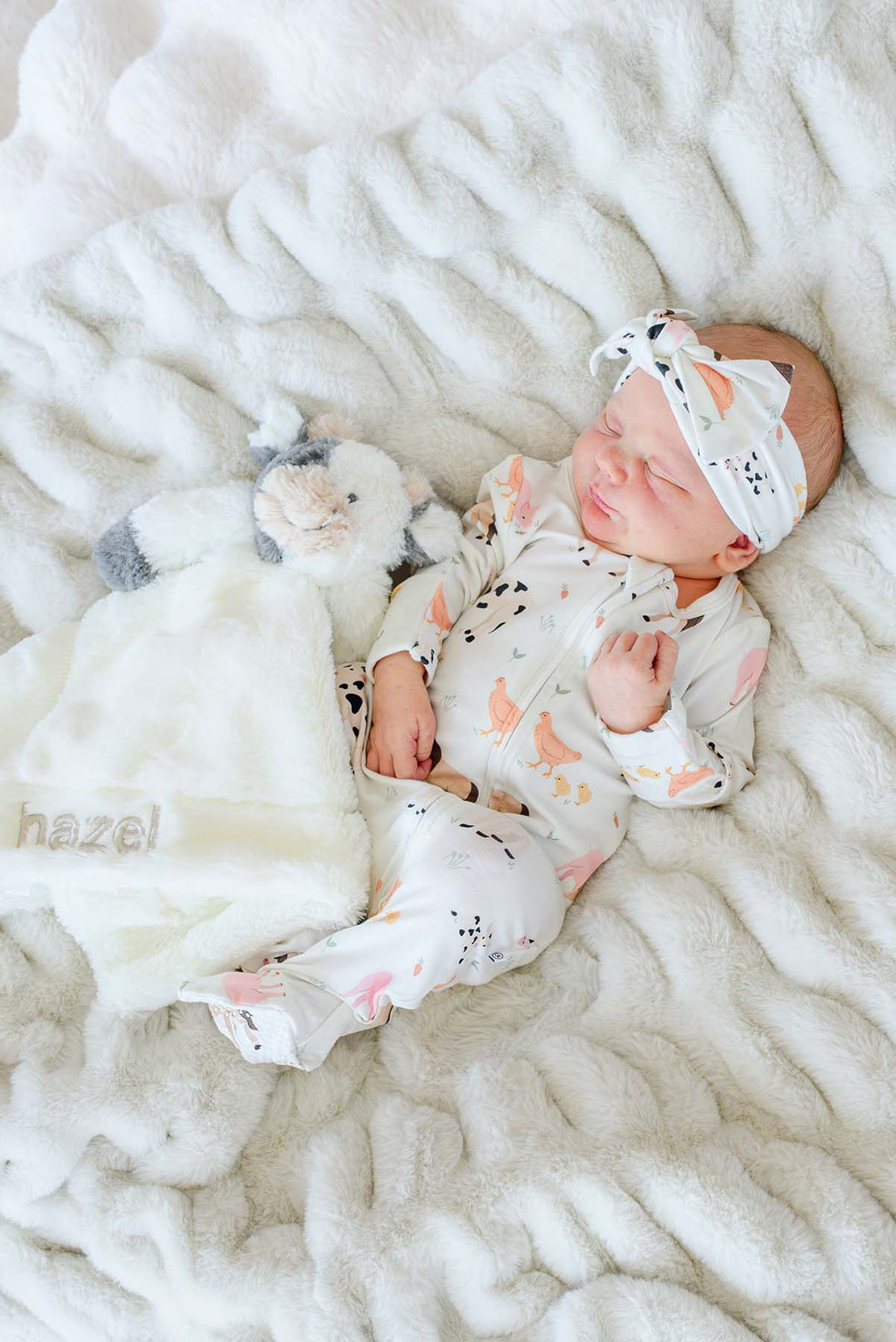 Sleeping newborn in Caden Lane farm animal footie pajamas and matching knotted headband, holding a pink faux fur lovey