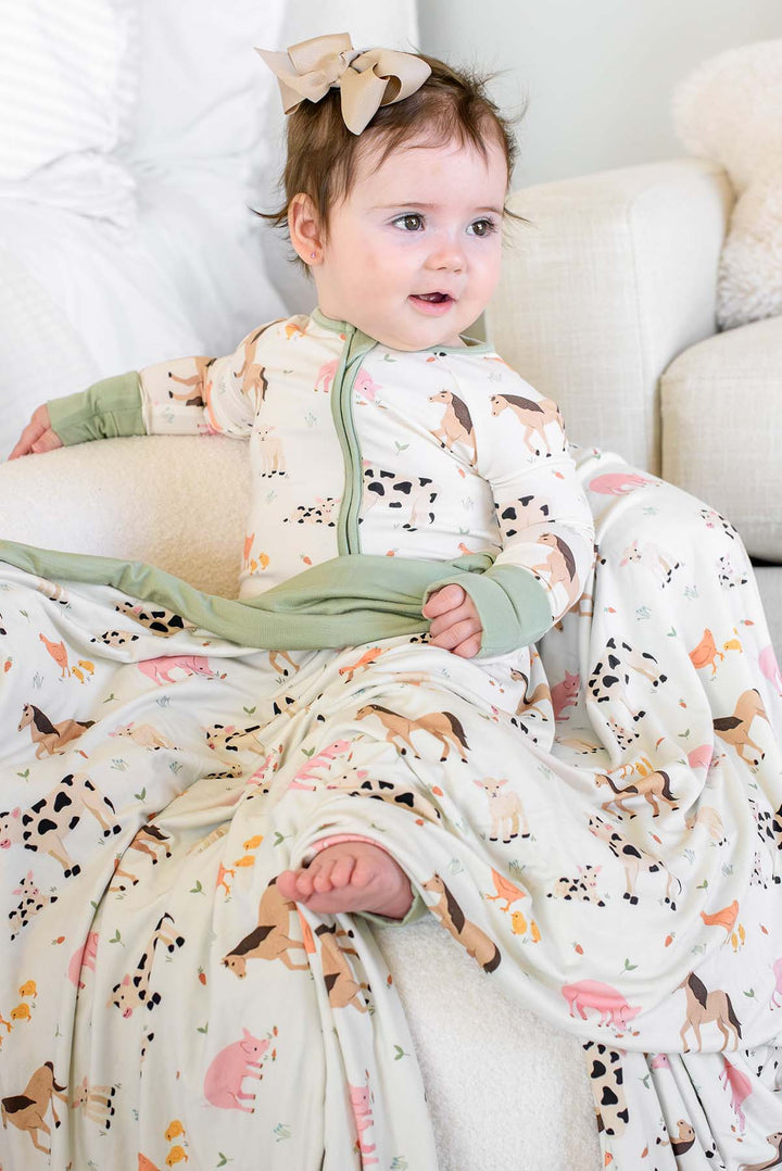 Baby girl in Caden Lane farm animal zip-up footie pajamas sitting with matching farm animal blanket with sage green trim