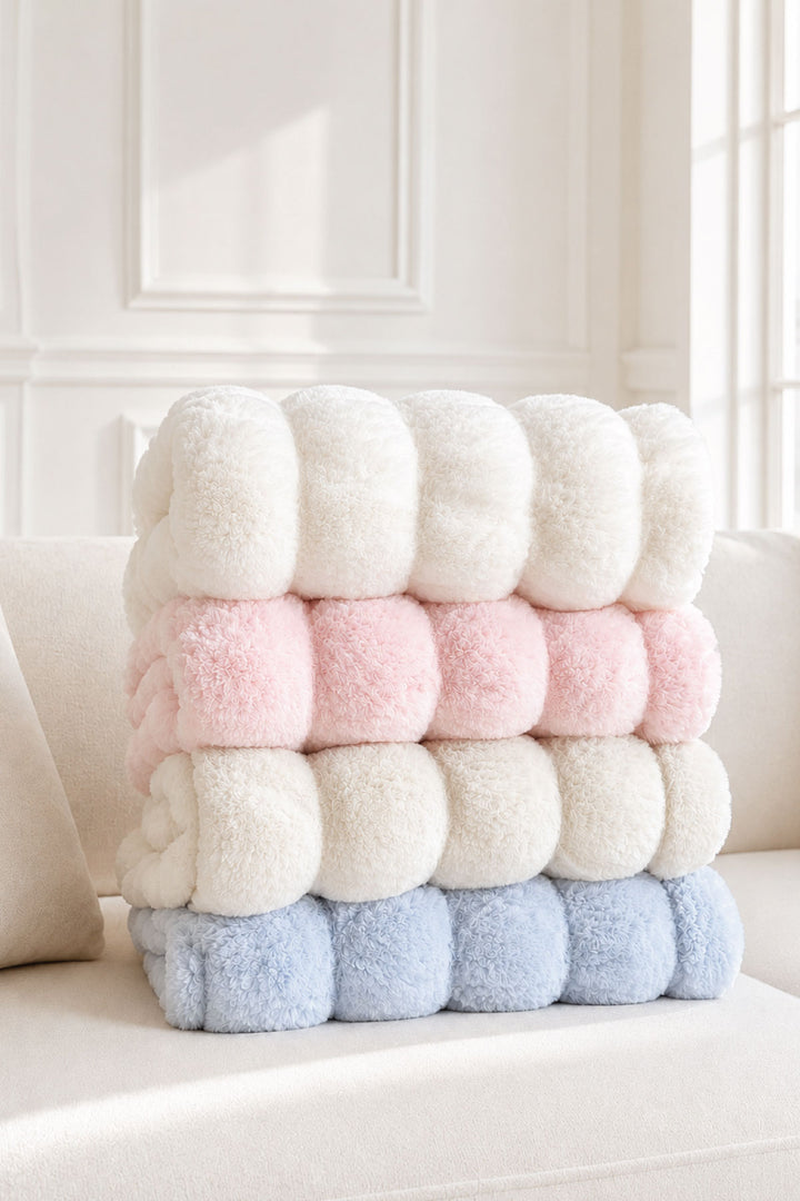 Stack of folded double-ruched faux fur baby blankets in cream, pink, cream, and blue layers on an armchair
