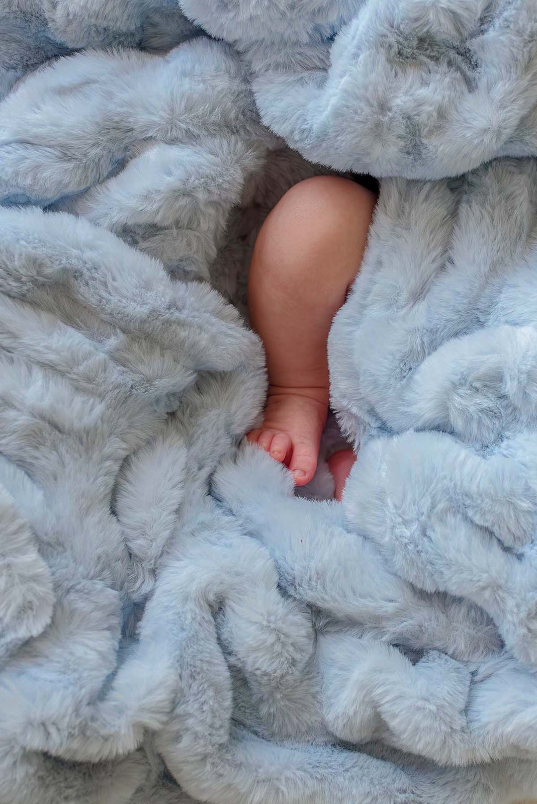 Close-up of a baby's leg nestled in the plush texture of an ice blue double-ruched faux fur blanket