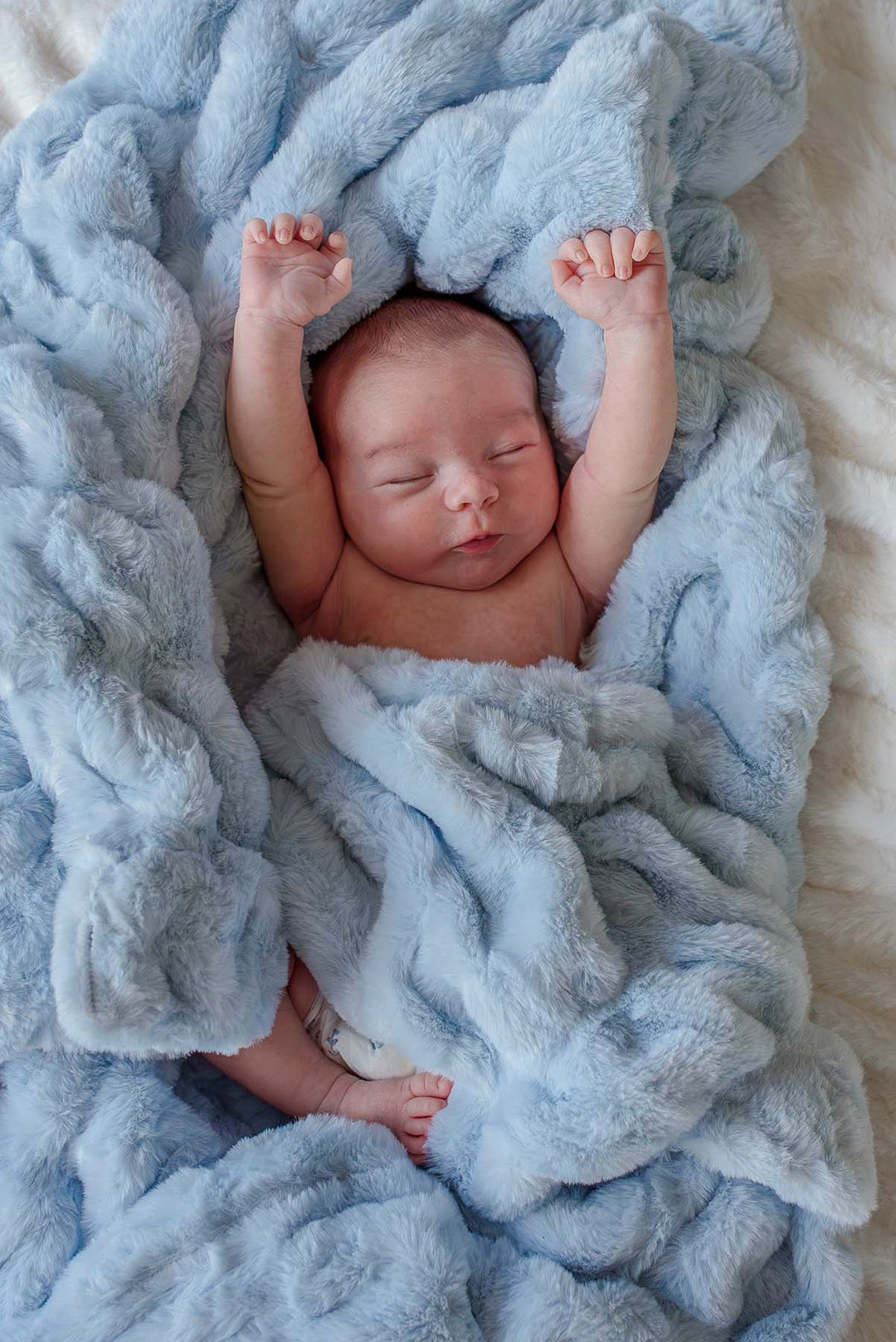 Newborn baby stretching with arms raised while lying on an ice blue double-ruched faux fur blanket