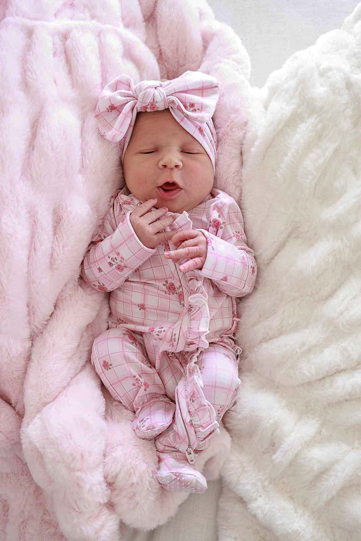 Newborn baby yawning in a pink plaid outfit and bow, lying on a barely pink double-ruched faux fur blanket