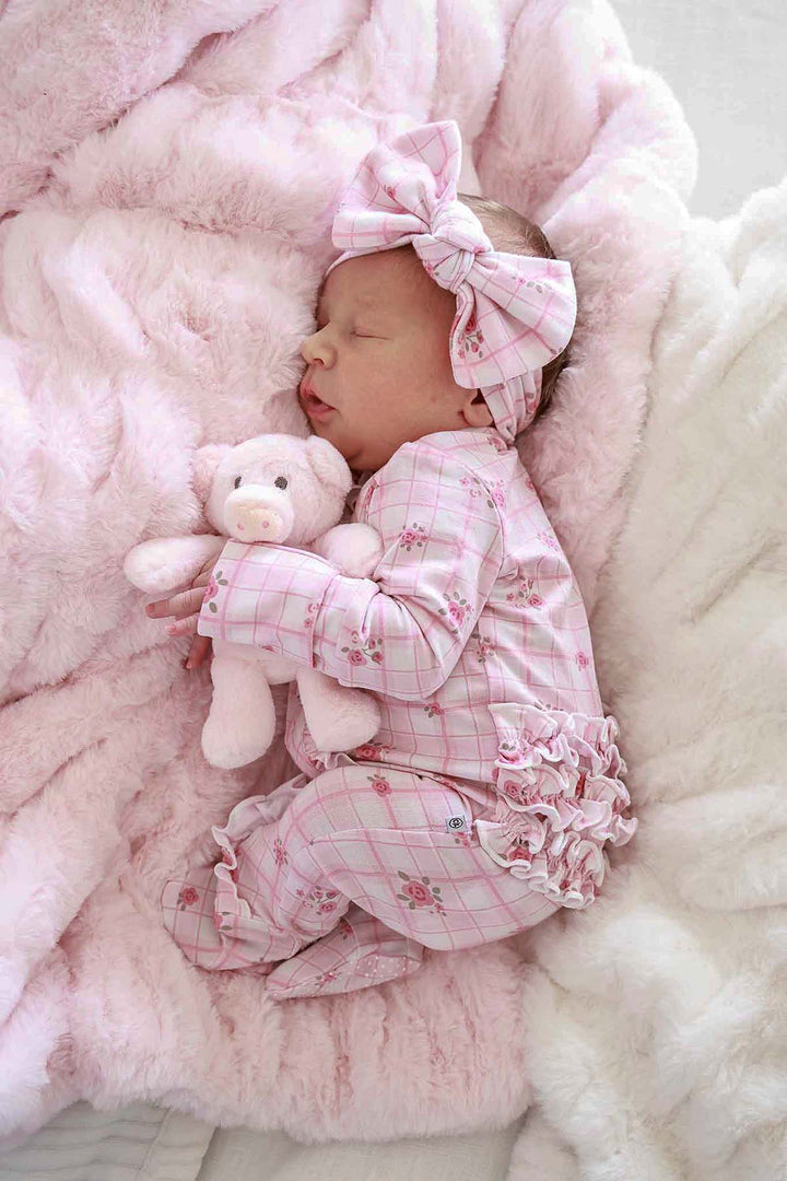 Sleeping baby in a pink plaid outfit and bow, holding a teddy bear on a barely pink double-ruched faux fur blanket