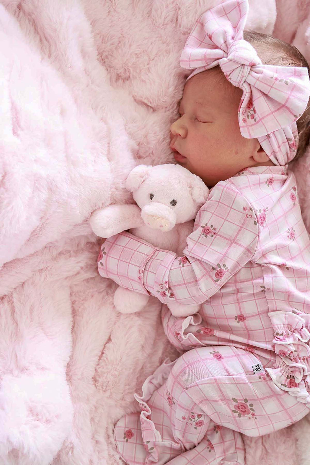 Close-up of sleeping baby in a pink plaid outfit hugging a teddy bear on a barely pink double-ruched faux fur blanket