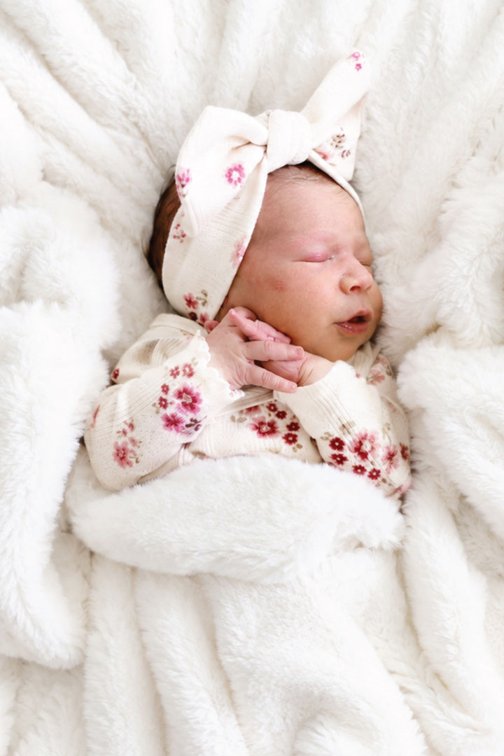 Sleeping newborn baby in a floral outfit and bow headband lying on a winter white double-ruched faux fur blanket