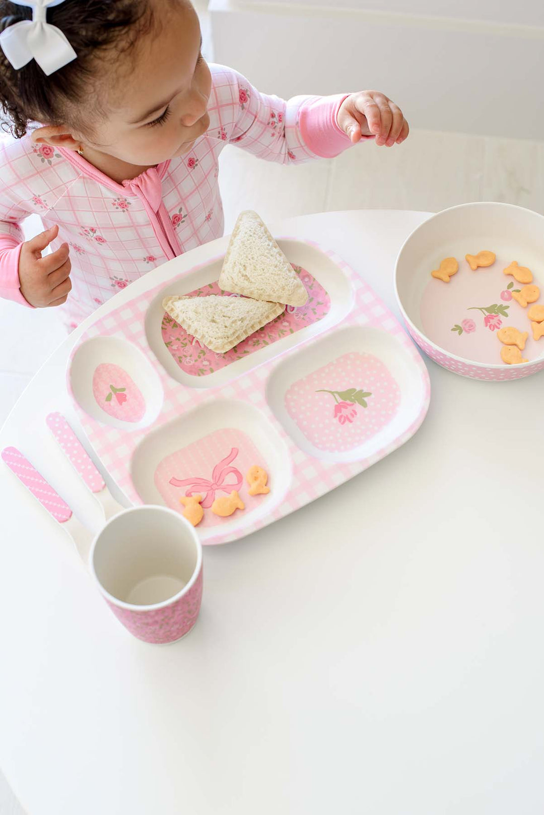 A young child wearing a 
Caden Lane pink plaid pajama top sits at a white table, using a Caden Lane pink and white gingham children's meal set. The set includes a compartment tray with rice balls and small orange snacks, a matching bowl, a cup, and a spoon.