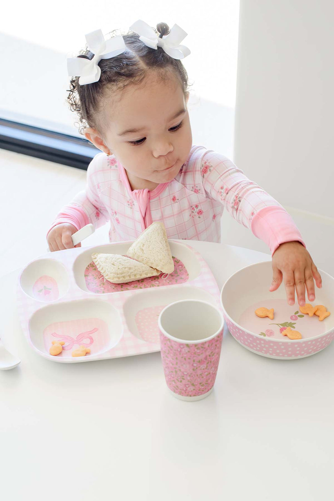 A toddler girl wearing a pink floral long-sleeve shirt and white bow headband sits at a white table, using a pink and white divided tray with a matching cup and bowl.