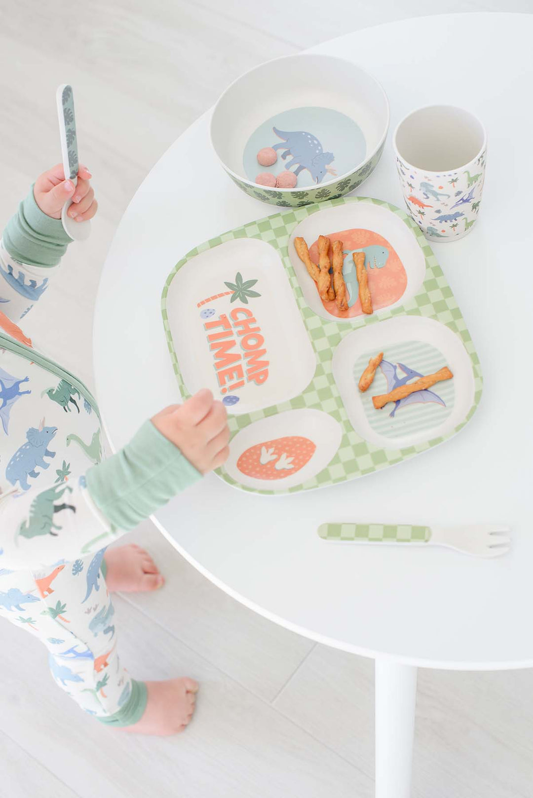 A child wearing dinosaur-print pajamas stands at a white table with a matching children's dinner set, including a divided plate, bowl, cup, and fork.